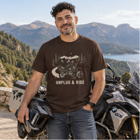 A man wearing a brown graphic t-shirt with 'UNPLUG & RIDE' printed on the front, standing outdoors next to a motorcycle with a mountain and lake landscape in the background.