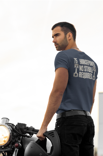 A man wearing a navy blue graphic t-shirt and black pants stands next to a motorcycle, holding a black helmet. The back of the t-shirt features white text that reads '75 HORSEPOWER NO STABLE REQUIRED.