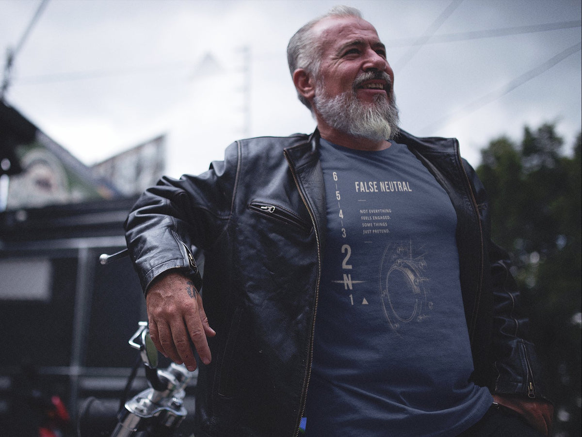 A smiling older man with a gray beard wearing a black leather motorcycle jacket over a dark blue graphic t-shirt, leaning against a motorcycle outdoors.