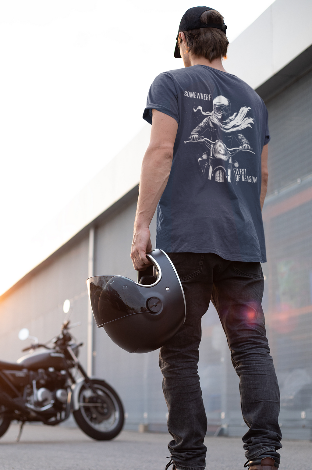 A man stands with his back to the camera, holding a black motorcycle helmet in one hand while wearing a navy blue graphic t-shirt, dark jeans, and brown boots. A black motorcycle is parked in the background near a modern building under bright sunlight.