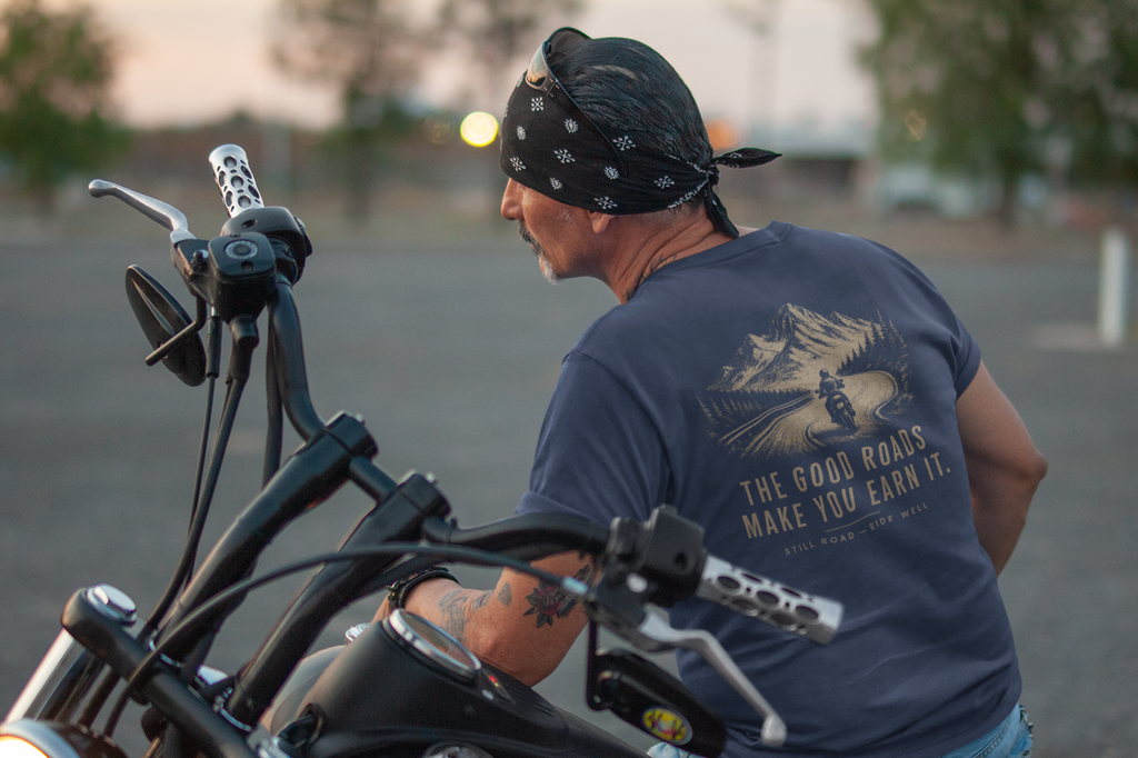 A man wearing a navy blue graphic t-shirt and a black bandana sits on a black cruiser motorcycle in an outdoor setting during sunset.