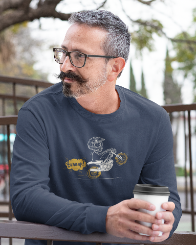 A man with glasses, a mustache, and a beard wearing a navy blue long-sleeve t-shirt with a cartoon graphic of a motorcycle and the word 'Braaap!'. He is holding a white paper coffee cup while leaning on a wooden railing in an outdoor setting.