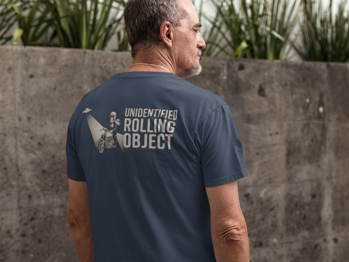 A man viewed from behind wearing a navy blue t-shirt with a white graphic print on the back that reads 'UNIDENTIFIED ROLLING OBJECT' next to a motorcycle illustration. He is standing outdoors in front of a textured concrete wall with green plants above it.