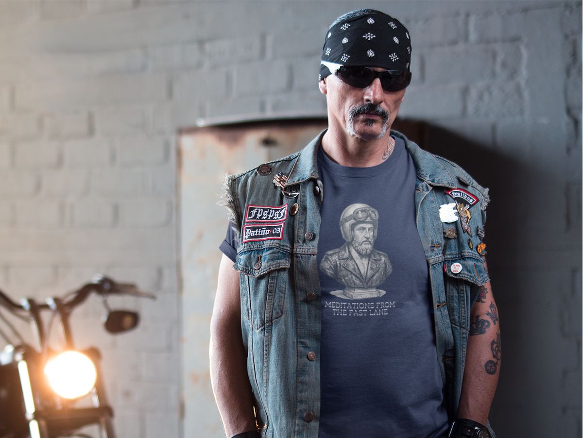 A man wearing a denim vest with patches, a graphic t-shirt, jeans, and a bandana stands next to a motorcycle in a garage setting.
