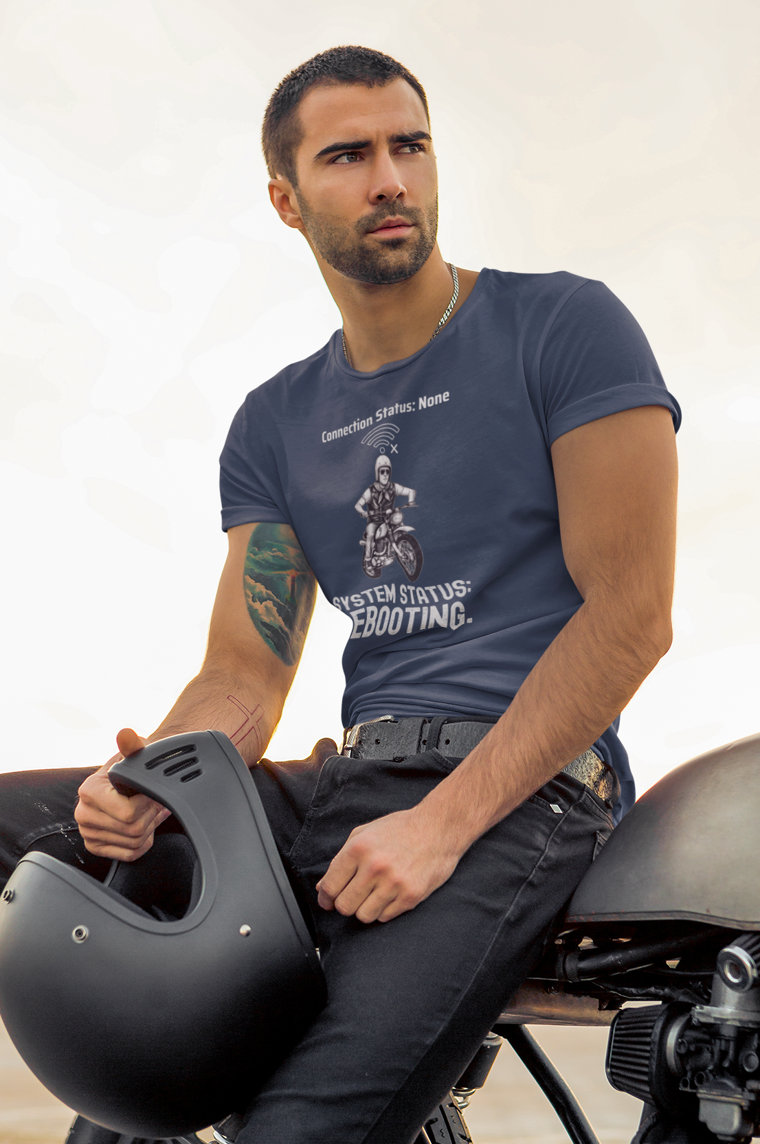 A man wearing a navy blue graphic t-shirt and black jeans sits on a motorcycle, holding a black helmet. The t-shirt features a white graphic of a motorcyclist with the text 'Rebooting'