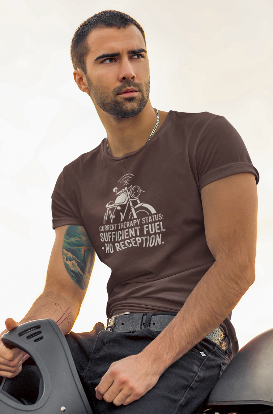 A man wearing a dark brown graphic t-shirt and black jeans sits on a motorcycle, holding a black helmet. The t-shirt features a white graphic of that says "No Reception" 