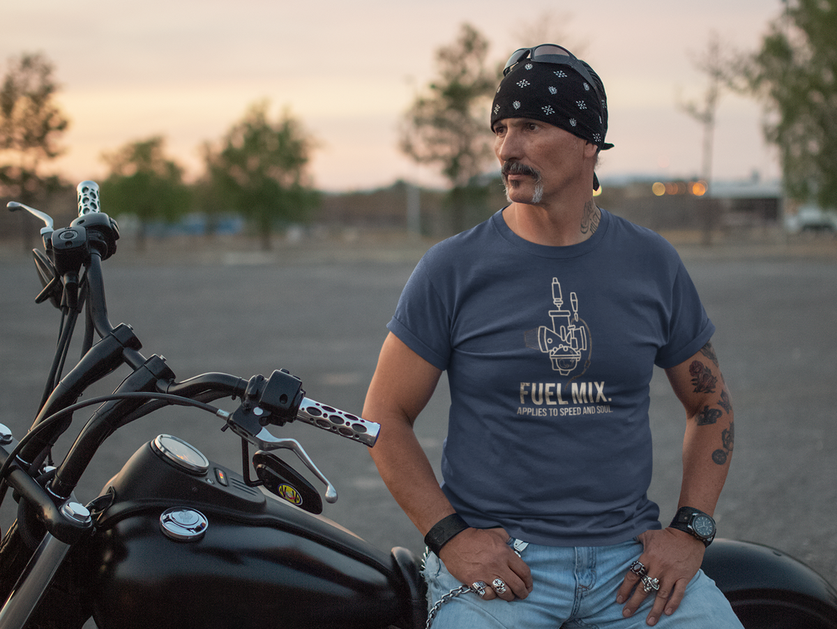 A man wearing a black bandana and a navy blue graphic t-shirt sits on a black cruiser motorcycle in an open paved lot at sunset. The t-shirt features a white graphic of a hand holding a cigarette with the text 'FUEL MIX'.