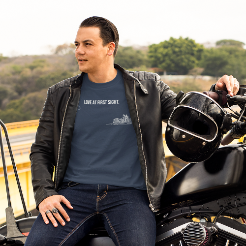 A man wearing a black leather jacket and a blue graphic t-shirt sits on a black cruiser motorcycle next to a yellow bridge with a hazy outdoor background.