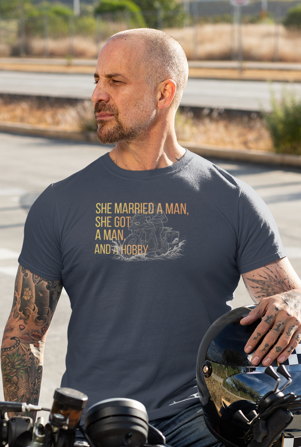 A man wearing a dark gray graphic t-shirt sits on a black motorcycle on an outdoor road. The t-shirt features a gold graphic of a woman and the text 'SHE MARRIED A MAN, SHE GOT A MAN, AND A HOBBY'.
