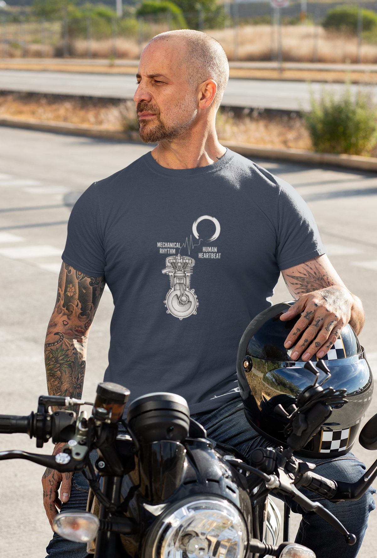 A man with tattoos sits on a motorcycle on a road, wearing a dark gray t-shirt with a white engine graphic and the text 'Mechanical Rhythm'. He is holding a black helmet.
