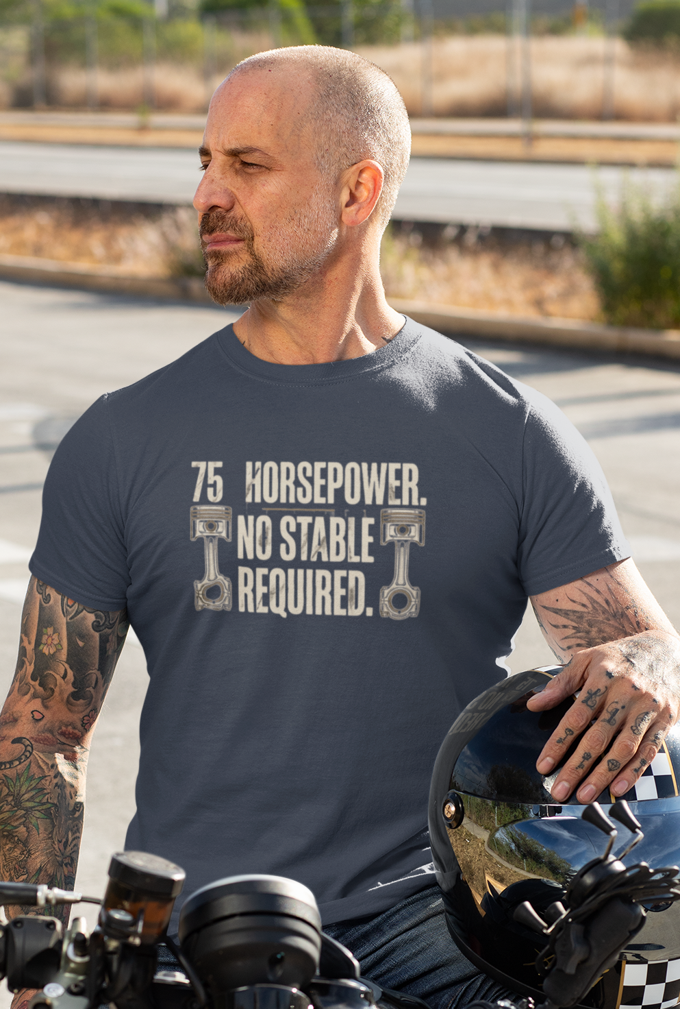 A man with tattoos sits on a motorcycle on a road, wearing a dark gray t-shirt with a white engine graphic and the text '75 Horsepower'. He is holding a black helmet.