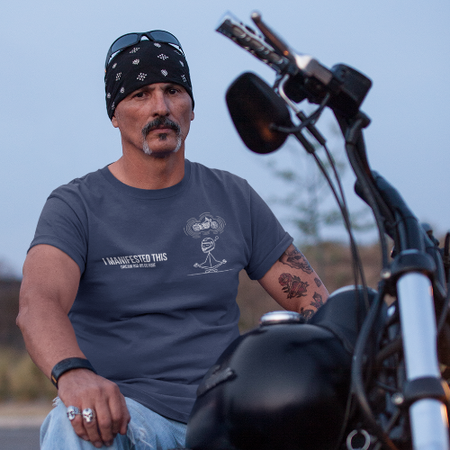 A man wearing a black bandana, a dark gray graphic t-shirt, and light blue jeans sits on a black cruiser motorcycle on an outdoor road during dusk.