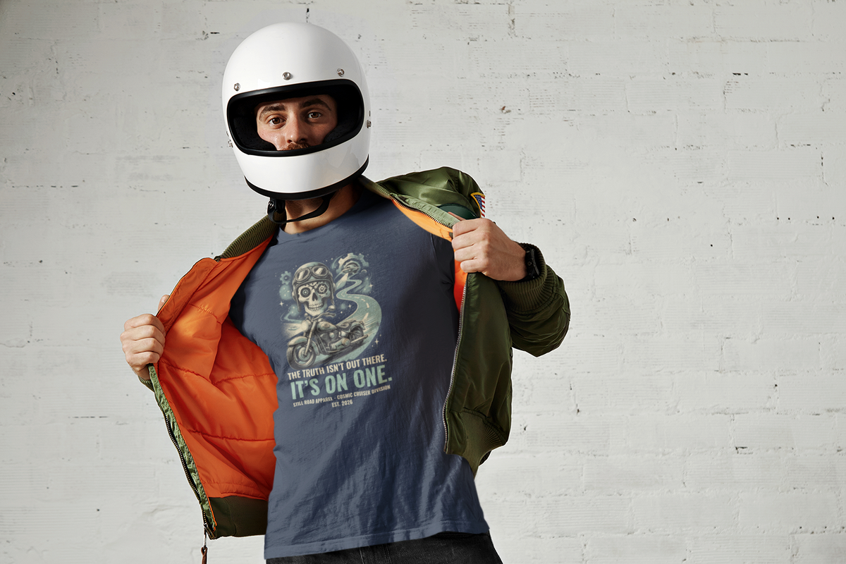 A man wearing a white motorcycle helmet and a blue graphic t-shirt stands against a white brick wall, opening an olive green jacket to reveal a bright orange interior.