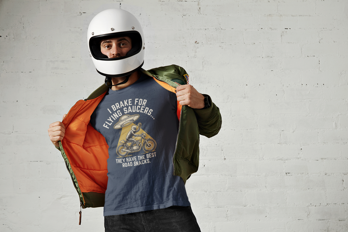 A man wearing a white motorcycle helmet and a blue graphic t-shirt stands against a white brick wall, opening an olive green jacket to reveal a bright orange lining. The t-shirt features a graphic of a motorcycle and the text 'I BRAKE FOR FLYING SAUCERS.