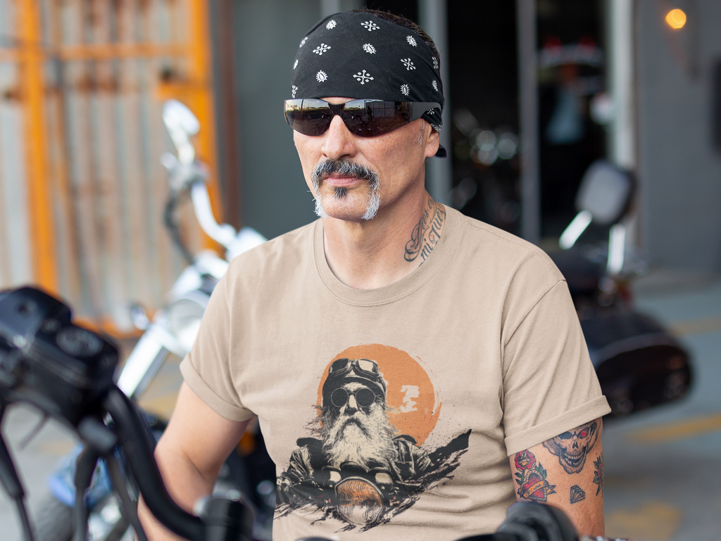 A man with a mustache and bandana sits on a motorcycle in front of a garage, wearing a beige graphic t-shirt with an OG biker graphic