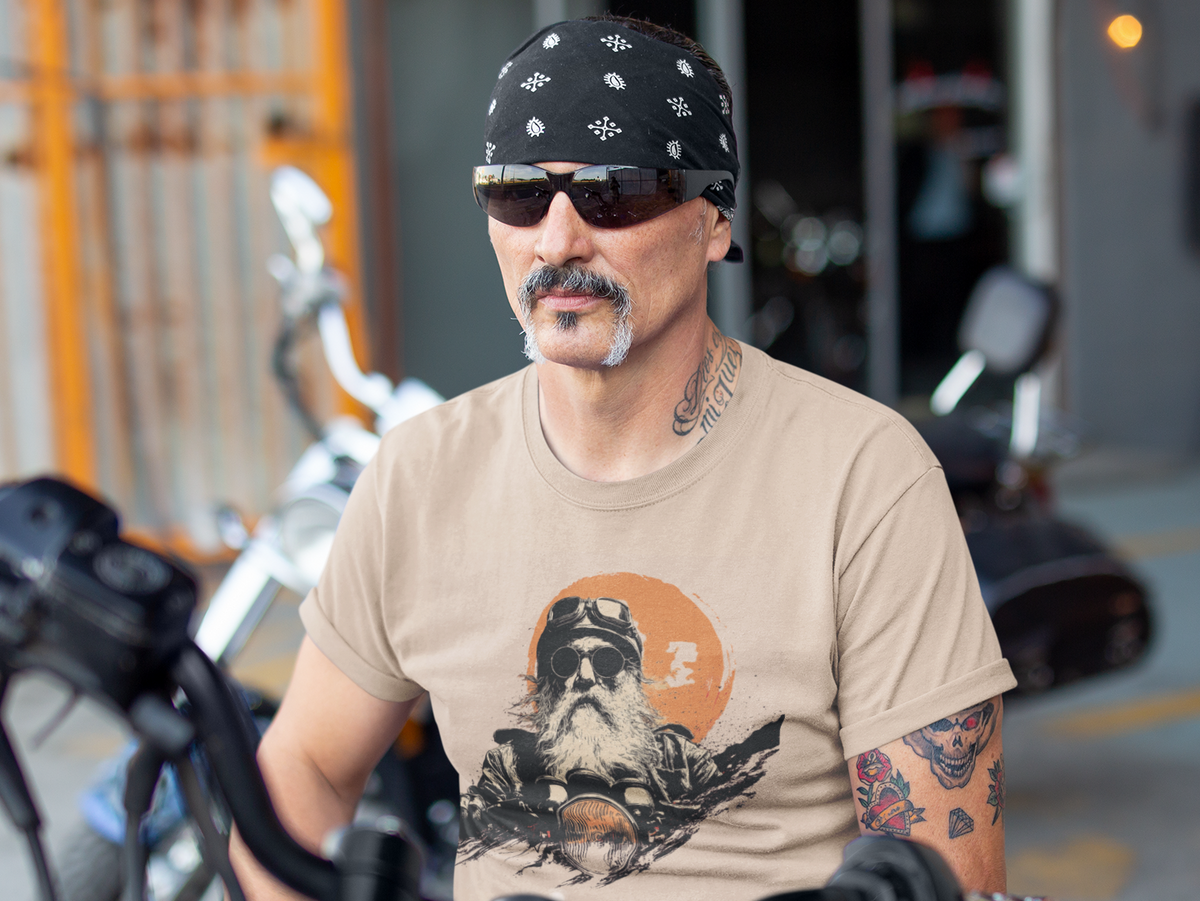 A man with a mustache and bandana sits on a motorcycle in front of a garage, wearing a beige graphic t-shirt with an OG biker graphic