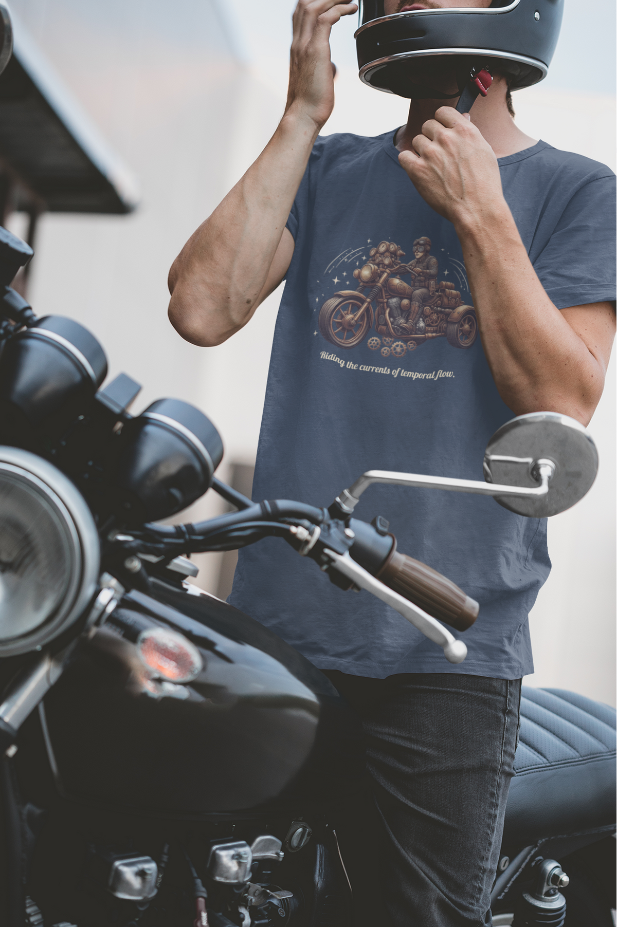 A man wearing a black motorcycle helmet and a navy blue graphic t-shirt stands next to a black cruiser motorcycle, adjusting his helmet.