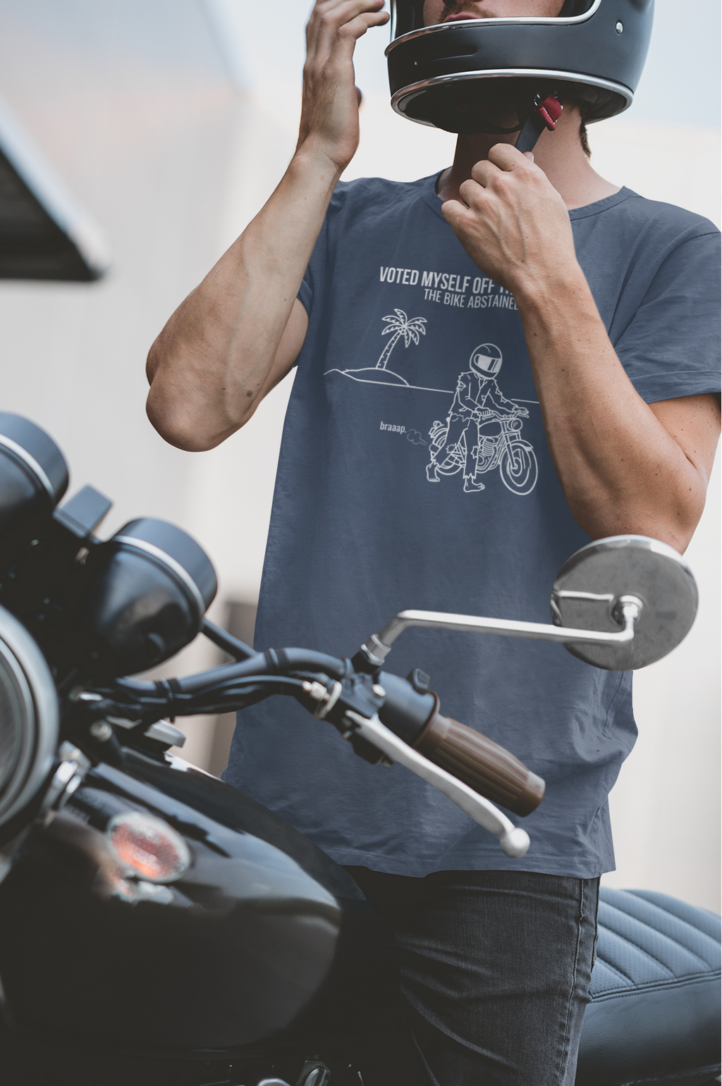 A man wearing a black motorcycle helmet and a navy blue graphic t-shirt stands next to a black cruiser motorcycle, adjusting his helmet.
