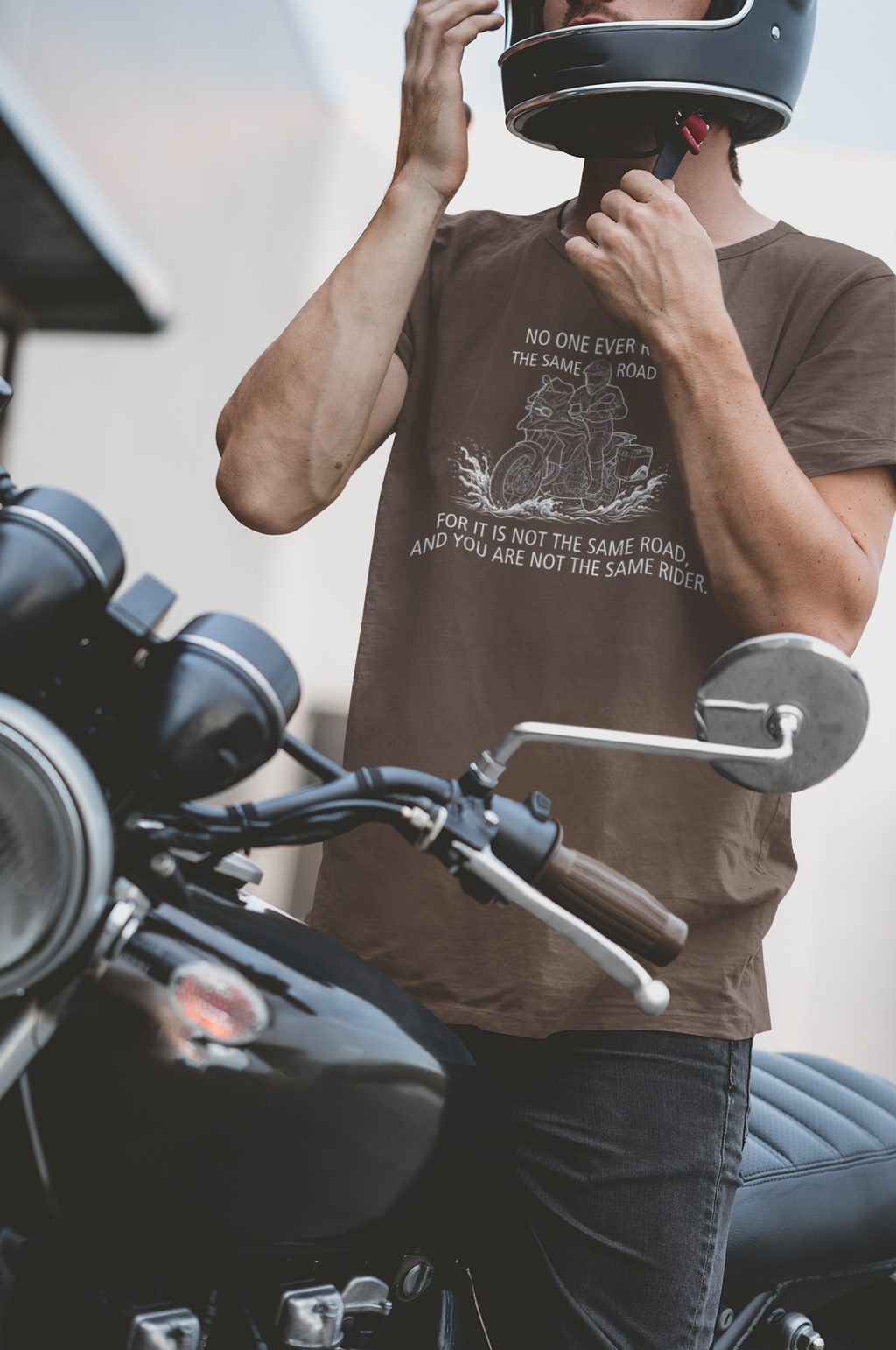 A man wearing a brown graphic t-shirt and a black motorcycle helmet sits on a black cruiser motorcycle, holding the helmet in place. The t-shirt features a white graphic of an ADV rider making a water crossing. The caption says 'NO ONE EVER RIDES THE SAME ROAD FOR IT IS NOT THE SAME ROAD AND YOU ARE NOT THE SAME RIDER'.