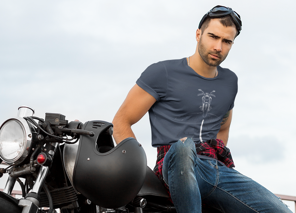 A man wearing a dark blue t-shirt, jeans, and red sneakers sits on a black cafe racer motorcycle on an outdoor bridge under a cloudy sky.