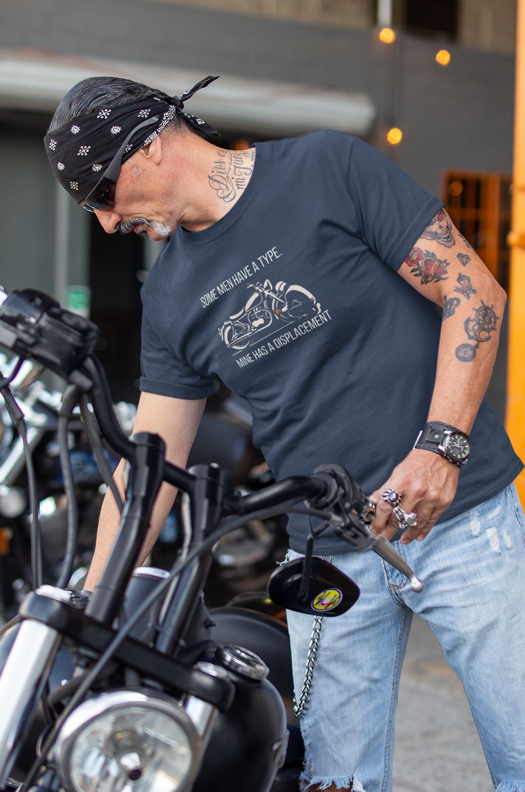 A man wearing a dark gray graphic t-shirt, ripped light blue jeans, and a black bandana leans over a black motorcycle in an industrial garage setting.