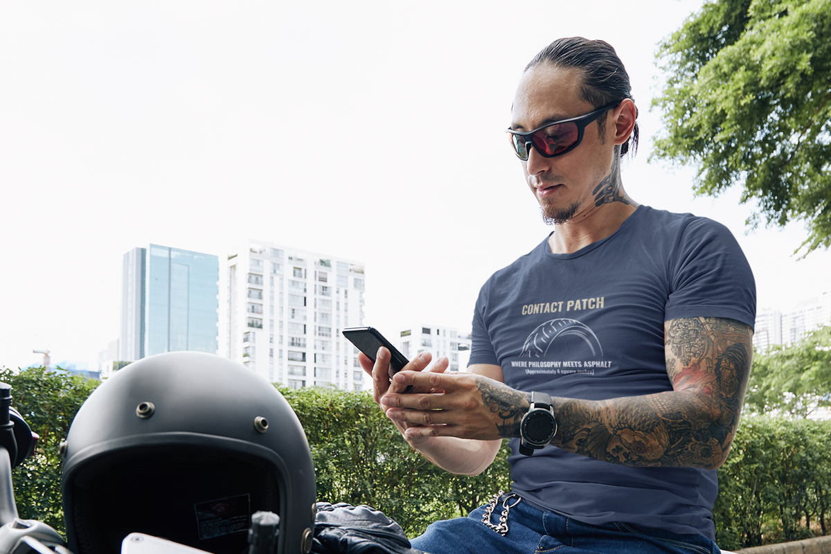 A man with tattoos and sunglasses sits on a motorcycle outdoors, looking at his phone while wearing a dark blue graphic t-shirt with 'CONTACT PATCH' printed on the front.