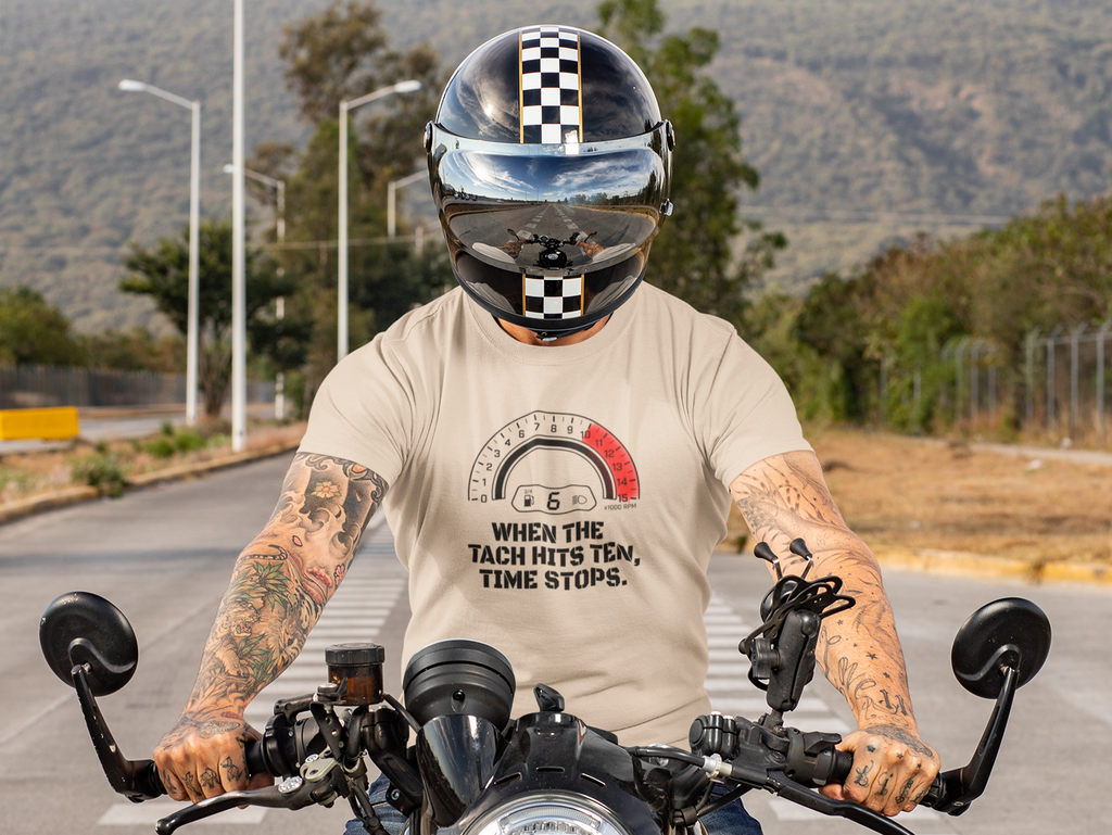 A tattooed man wearing a black and white checkered full-face helmet and a beige graphic t-shirt riding a black motorcycle on a paved road with hills in the background.
