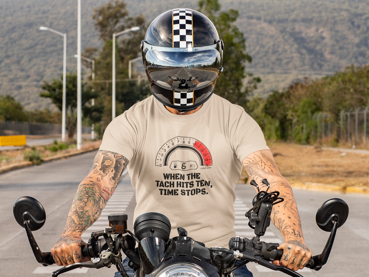 A tattooed man wearing a black and white checkered full-face helmet and a beige graphic t-shirt riding a black motorcycle on a paved road with hills in the background.