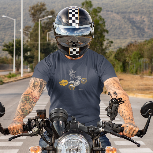 A tattooed man wearing a black and white checkered motorcycle helmet sits on a cruiser motorcycle on an outdoor road. He is wearing a dark gray t-shirt with a graphic print of a stick figure with a ridiculously large helmet riding a moto bike.