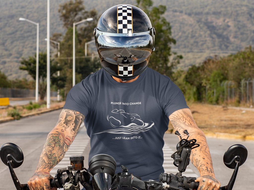 A tattooed man wearing a black and white checkered full-face helmet and a navy blue t-shirt with a white graphic print, riding a black motorcycle on a paved road with hills in the background.