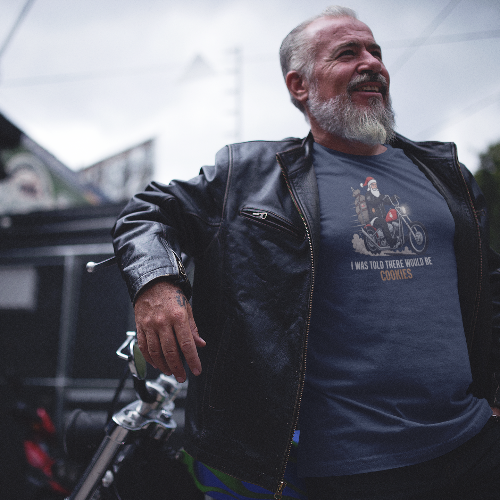 A smiling older man with a gray beard wearing a black leather jacket and a navy blue graphic t-shirt, leaning against a motorcycle outdoors.